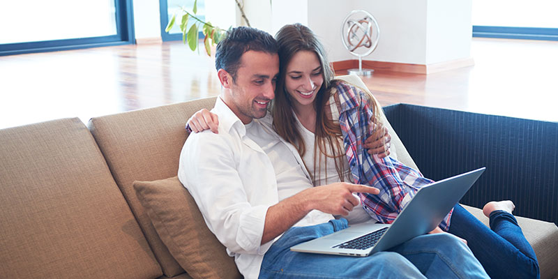 Smiling couple sitting on a couch, arm-in-arm, looking at a laptop in a bright living room with hardwood floors and large windows.