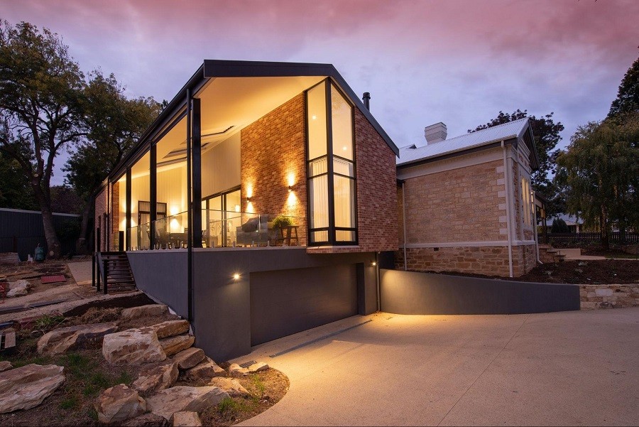 Photo of the front of a well-lit and beautifully landscaped brick home with large windows. 